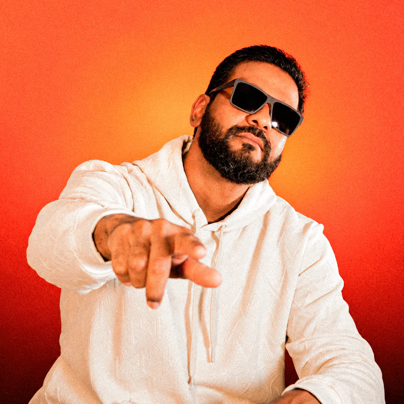 NuTom de óculos escuros e moletom branco, apontando pra frente, em fundo laranja